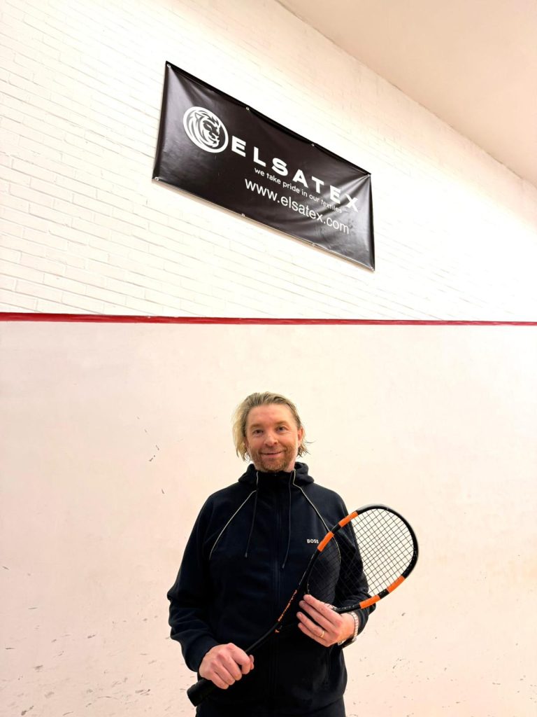 Elsatex CEO Stephen Lowe standing beneath the Official Partner banner at Mote Squash Club, holding his squash racket